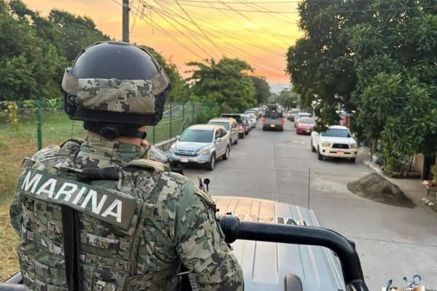 Mexican marine in fatigues standing in the bed of a pickup truck looking down an clear road with parked cars on either side