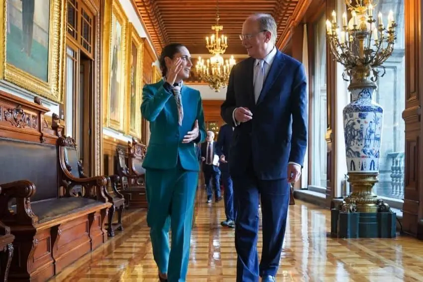 Mexican President Claudia Sheinbaum with Blackrock CEO Larry Fink in Mexico's National Palace. They are walking and talking to each other in a long elegant hallway with natural wood floors and paneling and a chandelier in the background