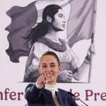 Mexico's President Claudia Sheinbaum at her daily press conference, pointing with her index finger straight into the camera as if taking a reporter's question. Behind her is the logo for her presidency, a black and white illustration of a young Mexican Indigenous woman in a traditional white Mexican dress and holding the Mexican flag.