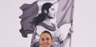 Mexico's President Claudia Sheinbaum at her daily press conference, pointing with her index finger straight into the camera as if taking a reporter's question. Behind her is the logo for her presidency, a black and white illustration of a young Mexican Indigenous woman in a traditional white Mexican dress and holding the Mexican flag.