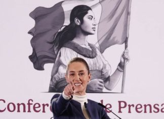 Mexico's President Claudia Sheinbaum at her daily press conference, pointing with her index finger straight into the camera as if taking a reporter's question. Behind her is the logo for her presidency, a black and white illustration of a young Mexican Indigenous woman in a traditional white Mexican dress and holding the Mexican flag.