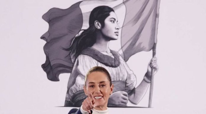 Mexico's President Claudia Sheinbaum at her daily press conference, pointing with her index finger straight into the camera as if taking a reporter's question. Behind her is the logo for her presidency, a black and white illustration of a young Mexican Indigenous woman in a traditional white Mexican dress and holding the Mexican flag.