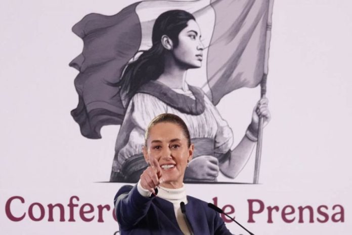 Mexico's President Claudia Sheinbaum at her daily press conference, pointing with her index finger straight into the camera as if taking a reporter's question. Behind her is the logo for her presidency, a black and white illustration of a young Mexican Indigenous woman in a traditional white Mexican dress and holding the Mexican flag.