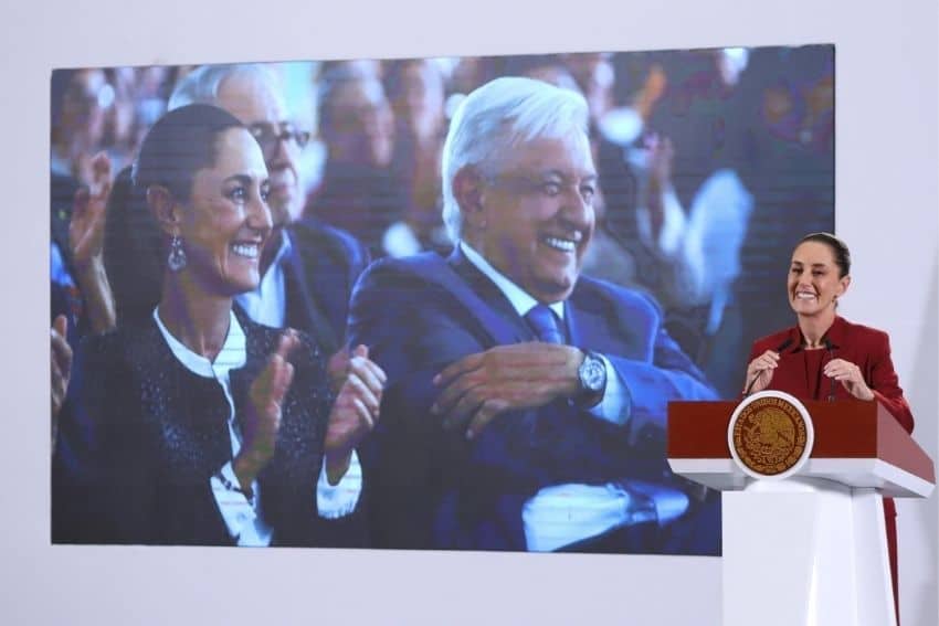 Claudia Sheinbaum at the presidential podium in front of a large projection of a photo of her and Andres Manuel Lopez Obrador smiling in the audience as they watch an event.