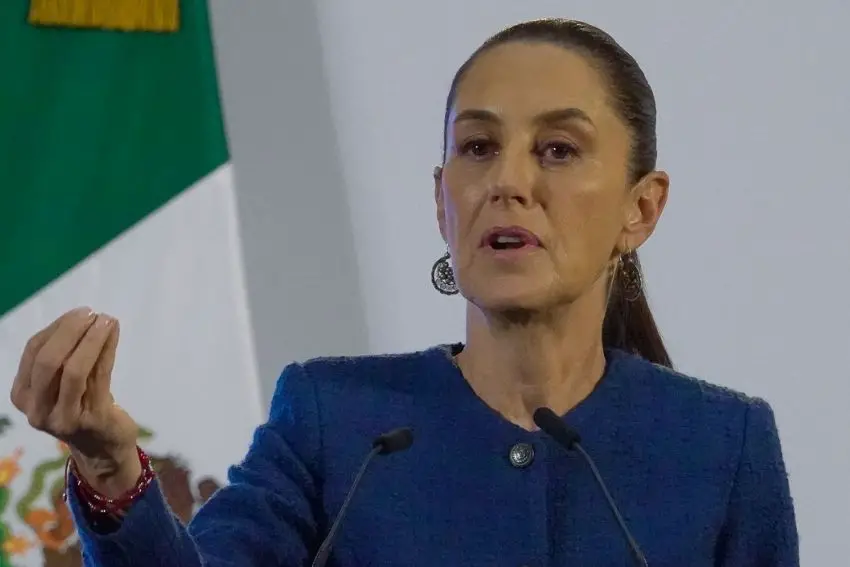 Claudia Sheinbaum in front of two small microphones at a press conference, gesturing with a single hand with her fingers closed together, in the middle of speaking