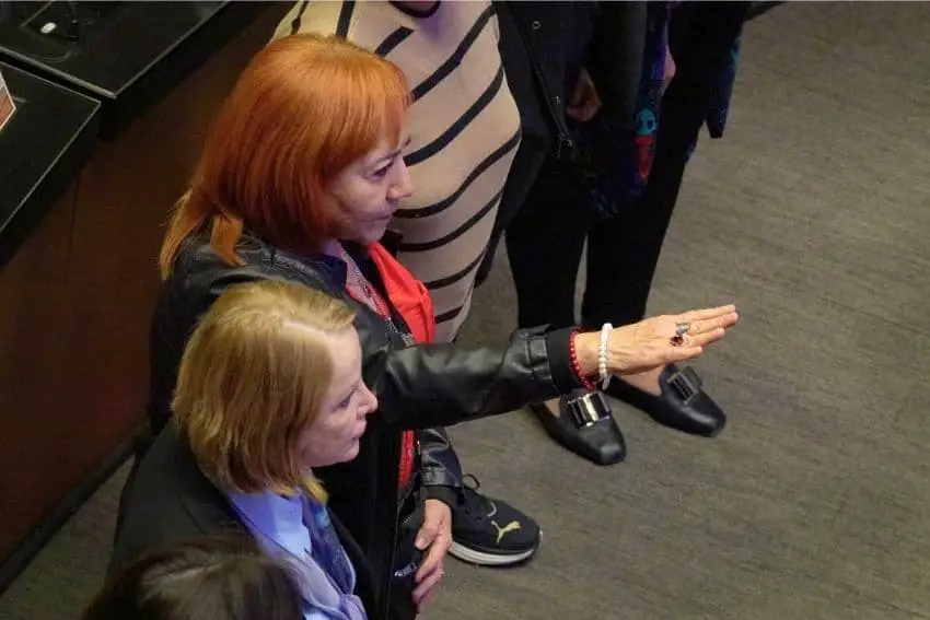 A red haired middle-aged Mexican woman in a black leather jacket holding her arm out formally as she is sworn into office on Mexico's senate floor. Next to her is another woman who is blonde and wearing a blue dress shirt. Both are looking forward to something off-camera.