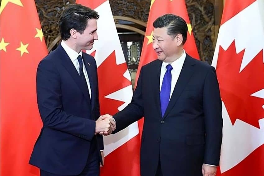 Justin Trudeau and Xi Jinping shaking hands in a formal photo shoot in front of Chinese and Canadian flags. They are smiling at each other.