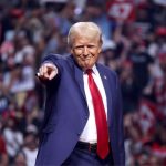 Donald Trump in a blue suit and red tie at a 2024 campaign rally, standing onstage in a stadium filled with people, smiling closemouthed as he points out into the audience