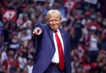 Donald Trump in a blue suit and red tie at a 2024 campaign rally, standing onstage in a stadium filled with people, smiling closemouthed as he points out into the audience