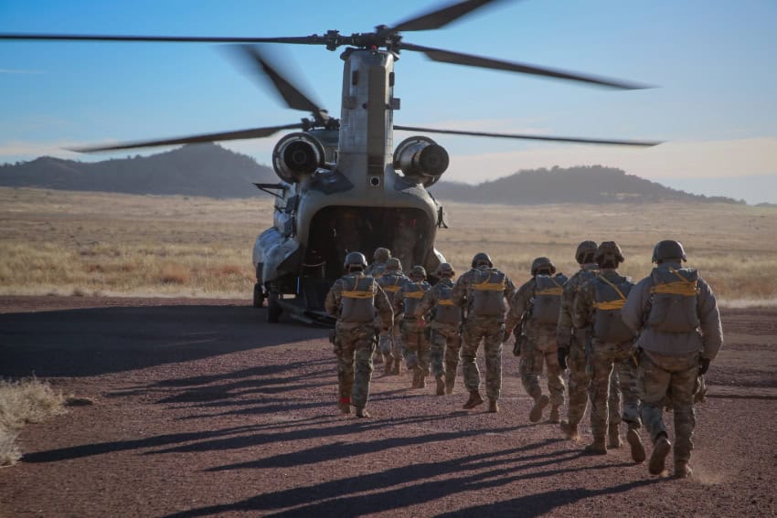 US troops at a military training event getting ready to board a military helicopter landed in front of a grassy plain with mountains in the background.