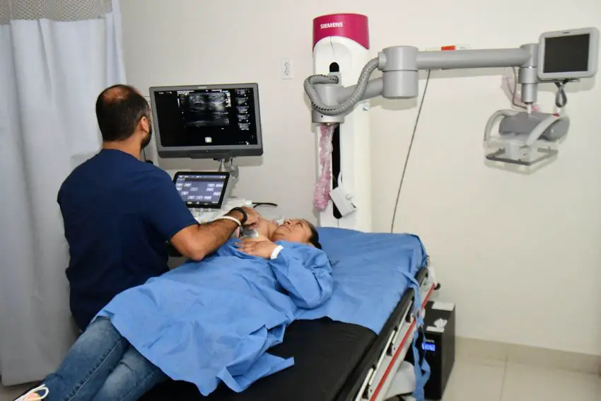 Mexican woman wearing a hospital gown and lying down on a hospital bed being given an ultrasound by a doctor who is looking at the ultrasound on a computer next to her bed.