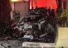 A mangled silver passenger vehicle after a car crash. It's located between a house and a large cement flowerpot from which can be seen growing tree trunks and green plants. Emergency officials are in the background behind the crashed vehicle.