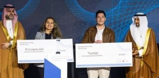 Male owner of Mexican company Yumari holding an oversized model of a bank check made out to the company for 100,000 US dollars standing next to a woman from the company Encapsulat holding up a check made out to Encapsulat for US $75,000. The pair are flanked at either end by two Saudi officials in traditional headdress and golden robes.