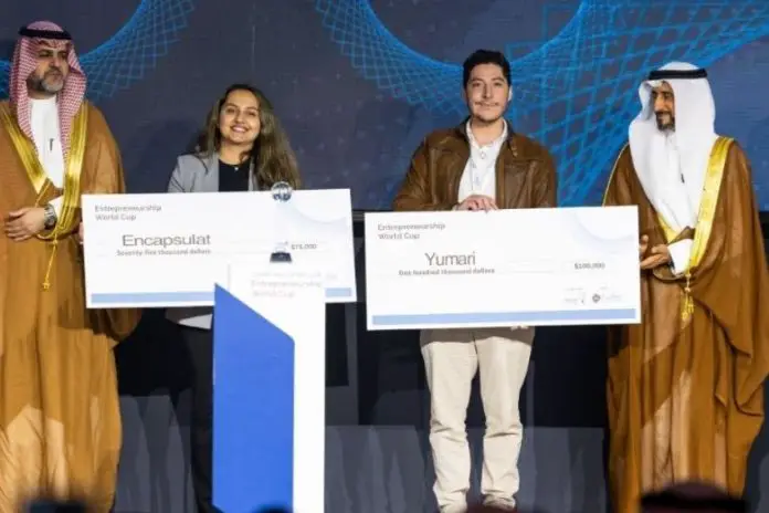 Male owner of Mexican company Yumari holding an oversized model of a bank check made out to the company for 100,000 US dollars standing next to a woman from the company Encapsulat holding up a check made out to Encapsulat for US $75,000. The pair are flanked at either end by two Saudi officials in traditional headdress and golden robes.