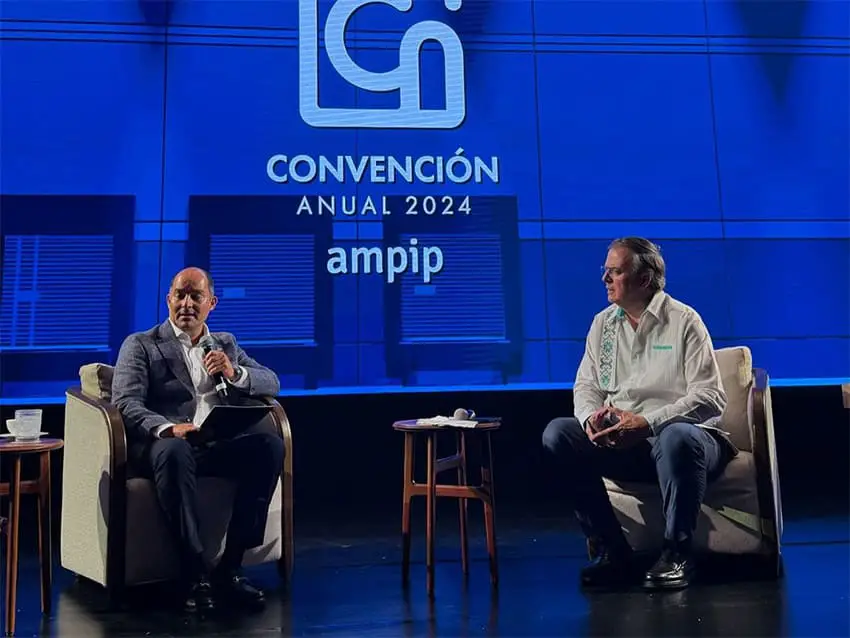 Marcelo Ebrard sits on a stage with another man, who is talking into a microphone