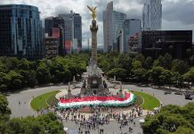 A crowd wraps Mexico City's Angel of Independence in a tricolored banner, with a view of the Mexico City skyline in the background