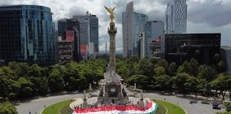 A crowd wraps Mexico City's Angel of Independence in a tricolored banner, with a view of the Mexico City skyline in the background