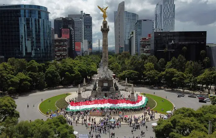 A crowd wraps Mexico City's Angel of Independence in a tricolored banner, with a view of the Mexico City skyline in the background