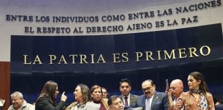 Senate President Gerardo Fernández Noroña, of the Morena party, sits on a legislative dias in front of the words "La Patria Es Primera" as lawmakers argue behind him.