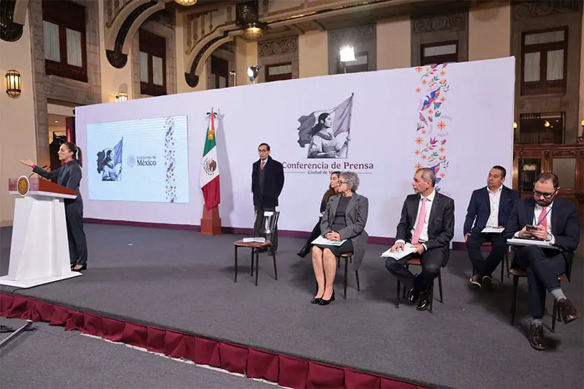 Sheinbaum stands at a podium on a stage, with government ministers and officials sitting behind her at her morning press conference.