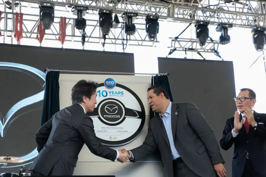 Gubernur Guanajuato Diego Sinhue berjabat tangan dengan pejabat Mazda Jepang di sebuah upacara. Di belakangnya ada tanda yang dibuat menyerupai roda penggerak mobil bertuliskan GTO, 10 tahun dan Mazda tipe besar.