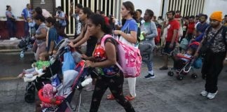 A dark-skinned young woman pushing a child in a stroller leads a group of migrants down a Mexican street, part of a migrant caravan