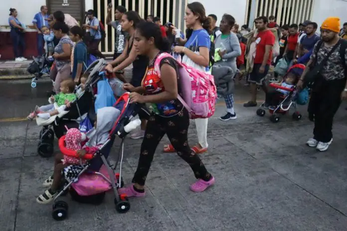 A dark-skinned young woman pushing a child in a stroller leads a group of migrants down a Mexican street, part of a migrant caravan