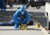 A forensic expert in blue protective gear and wearing a surgical mask and gloves in a city street in Chilpancingo puts down two yellow plastic marker signs on the ground to mark evidence in a crime scene. A dead victim lies sprawled on the sidewalk behind the expert.