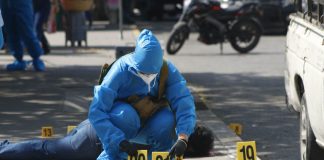 A forensic expert in blue protective gear and wearing a surgical mask and gloves in a city street in Chilpancingo puts down two yellow plastic marker signs on the ground to mark evidence in a crime scene. A dead victim lies sprawled on the sidewalk behind the expert.