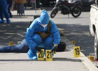 A forensic expert in blue protective gear and wearing a surgical mask and gloves in a city street in Chilpancingo puts down two yellow plastic marker signs on the ground to mark evidence in a crime scene. A dead victim lies sprawled on the sidewalk behind the expert.