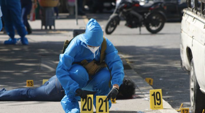 A forensic expert in blue protective gear and wearing a surgical mask and gloves in a city street in Chilpancingo puts down two yellow plastic marker signs on the ground to mark evidence in a crime scene. A dead victim lies sprawled on the sidewalk behind the expert.