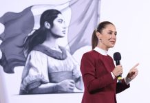 President Claudia Sheinbaum holding a microphone in front of her administration's logo of a Indigenous Mexican woman in front of the Mexican flag.