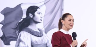 President Claudia Sheinbaum holding a microphone in front of her administration's logo of a Indigenous Mexican woman in front of the Mexican flag.