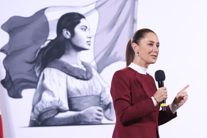 President Claudia Sheinbaum holding a microphone in front of her administration's logo of a Indigenous Mexican woman in front of the Mexican flag.