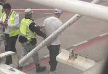 Outside a Volaris airplane window, a Mexican National Guard member in a yellow safety vest escorts a handcuffed man in a white T-shirt away from the plane.