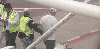 Outside a Volaris airplane window, a Mexican National Guard member in a yellow safety vest escorts a handcuffed man in a white T-shirt away from the plane.