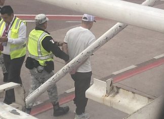 Outside a Volaris airplane window, a Mexican National Guard member in a yellow safety vest escorts a handcuffed man in a white T-shirt away from the plane.