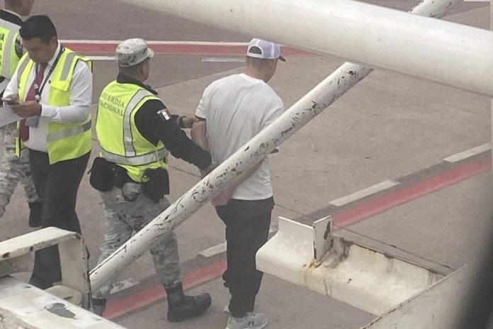 Outside a Volaris airplane window, a Mexican National Guard member in a yellow safety vest escorts a handcuffed man in a white T-shirt away from the plane.