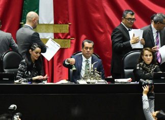 Mexican Federal Deputy Sergio Gutiérrez and head of the board of directors of the Chamber of Deputies rings a bell in to open session. He's sitting at a desk at the head of the Chamber with other members of the board of directors sitting on either side of him and other lawmakers standing behind them, conducting other business