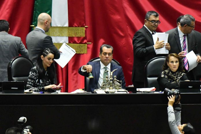 Mexican Federal Deputy Sergio Gutiérrez and head of the board of directors of the Chamber of Deputies rings a bell in to open session. He's sitting at a desk at the head of the Chamber with other members of the board of directors sitting on either side of him and other lawmakers standing behind them, conducting other business