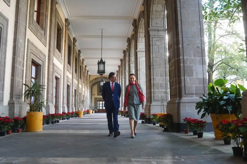 Mexico's President Claudia Sheinbaum and Colombia's President Gustavo Preto walking down a long courtyard of Mexico's National Palace toward the camera. Around them are windows to the building on one side and massive arched pillars on the other. Both sides of the courtyard are decorated with plants, including many poinsettias.