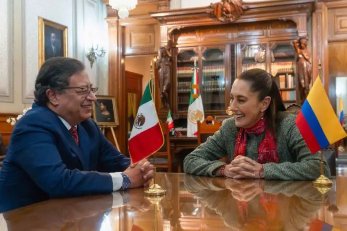 Colombian President Gustavo Petro sits across from Mexican President Claudia Sheinbaum in a room in Mexico's National Palace. Each have next to them small brass stands holding a small flag of each other's country. They are smiling and in mid conversation.