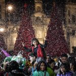 A child sits on an adults shoulders at the Mexico City Christmas Verbena, with giant Christmas trees in the background and fake snow falling