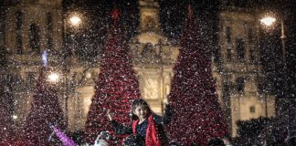 A child sits on an adults shoulders at the Mexico City Christmas Verbena, with giant Christmas trees in the background and fake snow falling