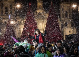 A child sits on an adults shoulders at the Mexico City Christmas Verbena, with giant Christmas trees in the background and fake snow falling