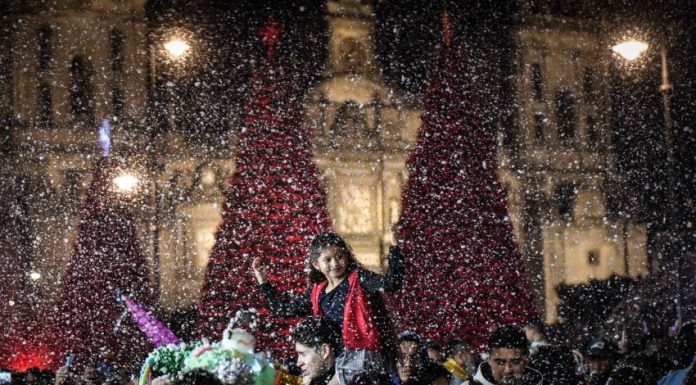 A child sits on an adults shoulders at the Mexico City Christmas Verbena, with giant Christmas trees in the background and fake snow falling