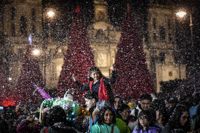 A child sits on an adults shoulders at the Mexico City Christmas Verbena, with giant Christmas trees in the background and fake snow falling