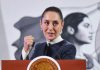 Claudia Sheinbaum in mid speech while standing at the presidential podium during a press conference. She is looking forward with her gaze pointed toward her left and is holding her right hand up before her chest in a closed fist.