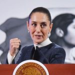 Claudia Sheinbaum in mid speech while standing at the presidential podium during a press conference. She is looking forward with her gaze pointed toward her left and is holding her right hand up before her chest in a closed fist.