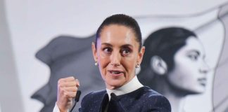 Claudia Sheinbaum in mid speech while standing at the presidential podium during a press conference. She is looking forward with her gaze pointed toward her left and is holding her right hand up before her chest in a closed fist.